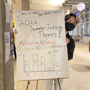 Photo of Isaac posing with a poster of the 2022 Summer Institute theme voting results.