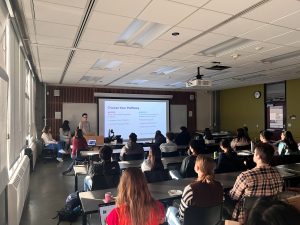 A photo of someone presenting in front of a screen. The office of undergrad research presentation with students sitting and watching.