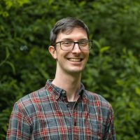 Headshot of Keenan. Keenan smiling for the photo in front of greenery