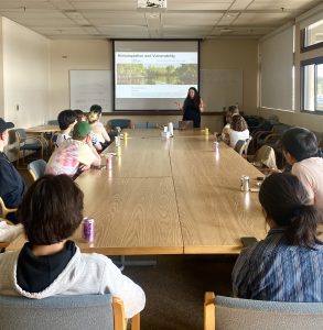 Dr. Valentine presenting on blue carbon in front of a screen. Students are seen sitting around a long table watching.