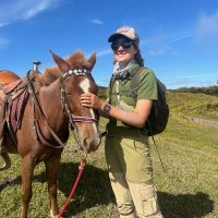 Photo of Ellie outdoors, standing with a horse, and smiling