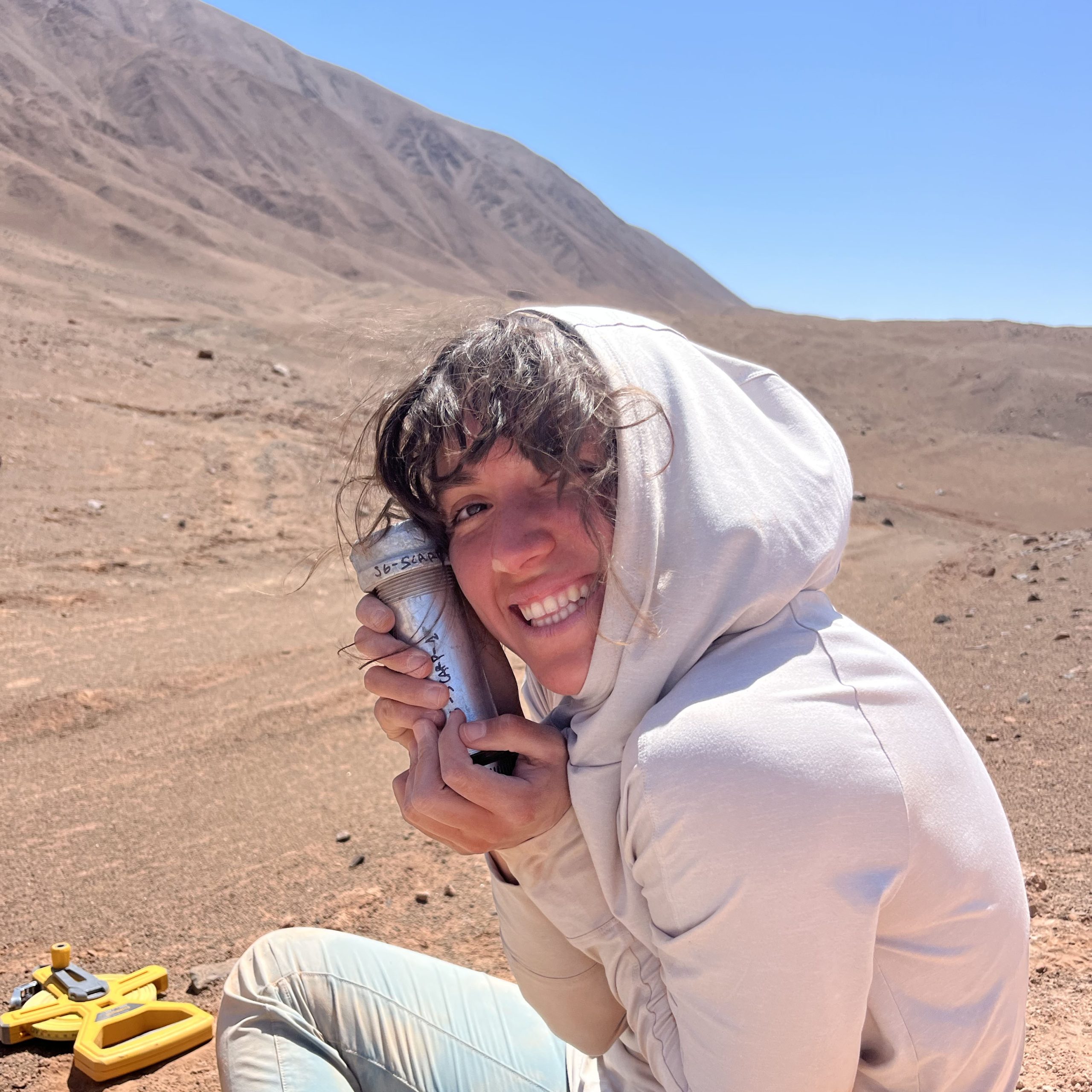 Tamara in the desert sitting down looking over shoulder smiling with her hood on holding sampling supplies